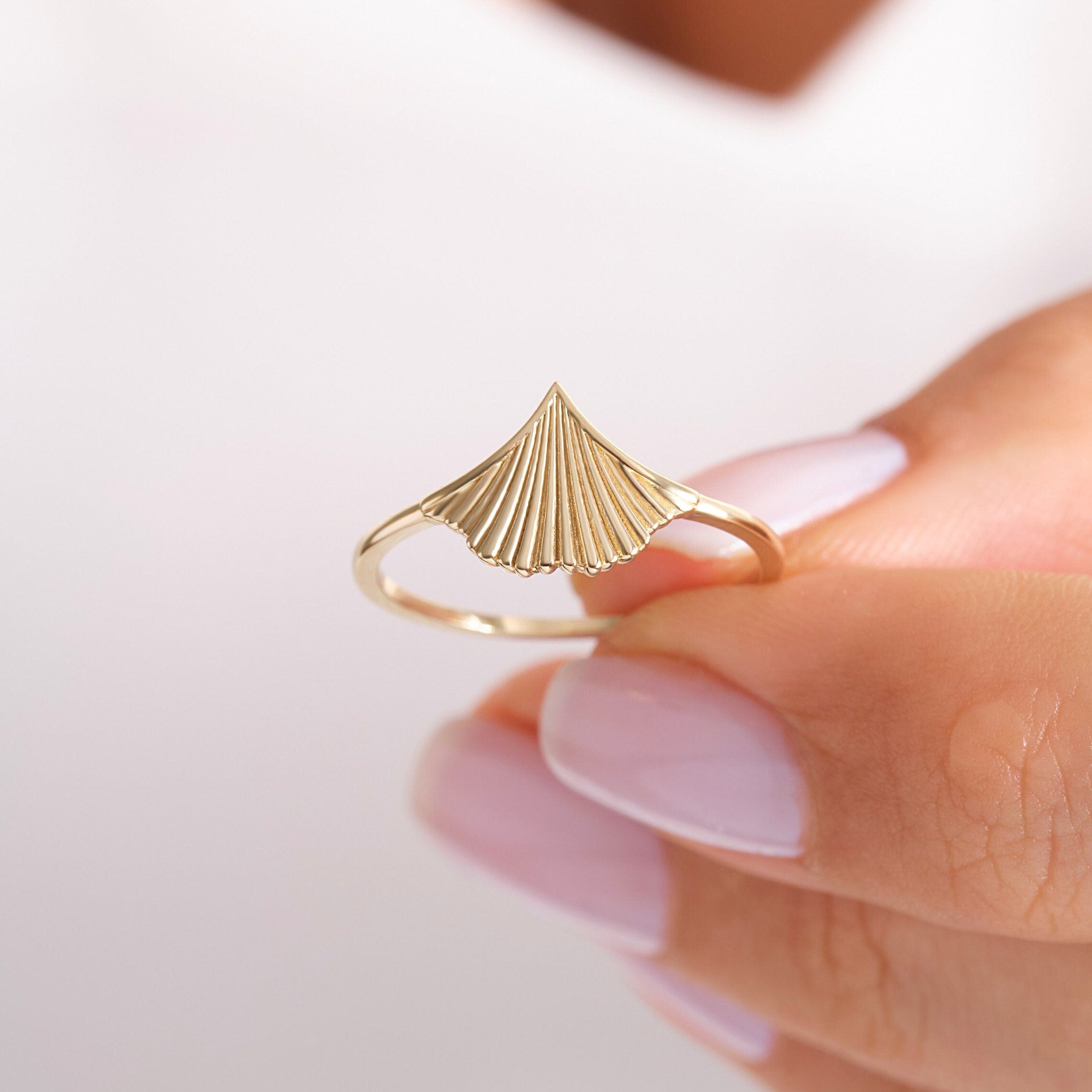 May include: A gold ring features a unique fan-shaped design. The ring's band is thin and delicate, while the centerpiece is a striking, textured fan shape that adds a touch of elegance. The fan-shaped design is meticulously detailed, with each line and curve adding to its overall appeal. This gold ring is a statement piece that would make a perfect addition to any jewelry collection.
